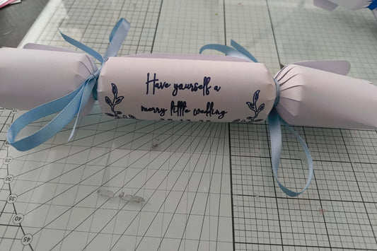 White cracker with navy foil wedding lettering “Have yourself a merry little wedding,” tied with pale blue ribbon on craft background.