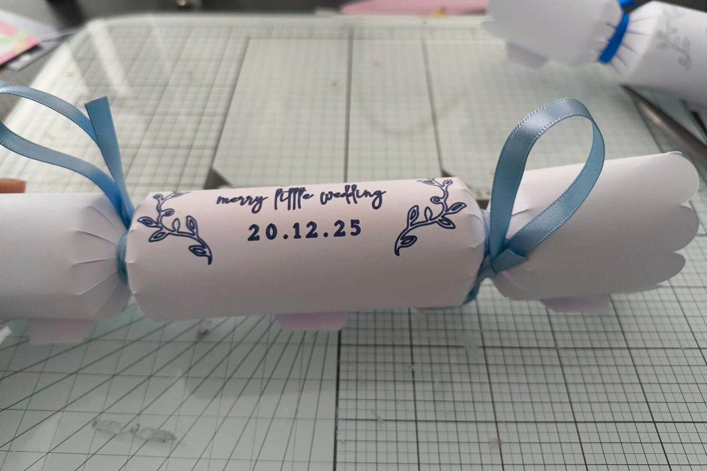 White cracker with navy foil wedding lettering and date, finished with pale blue ribbons, styled flat on craft mat background.