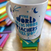 A white ceramic mug with blue text that reads: "THE WORLD NEEDS MORE TEACHERS LIKE YOU." Surrounding the text are blue decorative elements, including stars, a crescent moon, and abstract shapes resembling shooting stars. The mug has a handle on the left and rests on a neutral surface, radiating a sense of appreciation for educators.