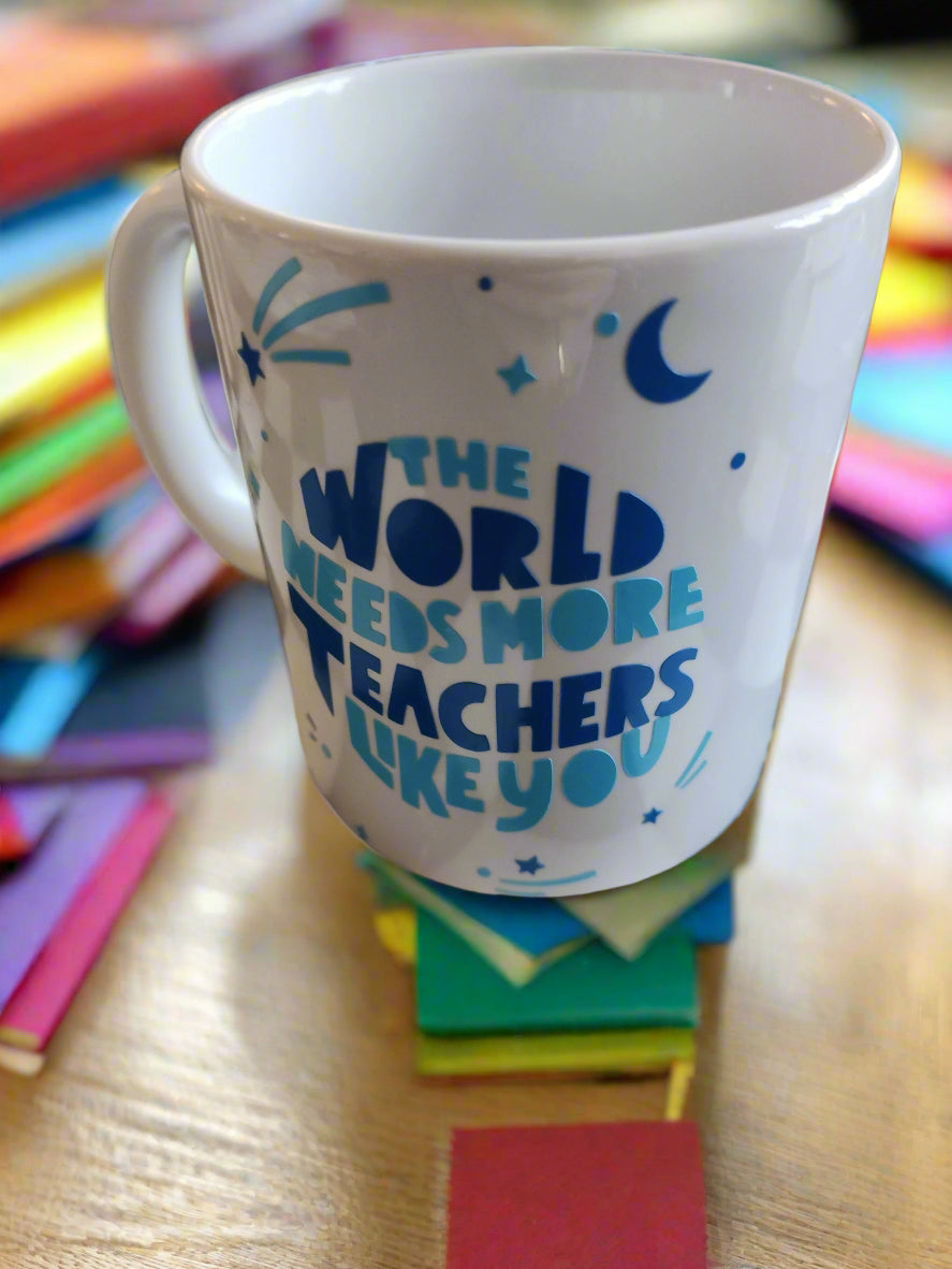 A white ceramic mug with blue text that reads: "THE WORLD NEEDS MORE TEACHERS LIKE YOU." Surrounding the text are blue decorative elements, including stars, a crescent moon, and abstract shapes resembling shooting stars. The mug has a handle on the left and rests on a neutral surface, radiating a sense of appreciation for educators.