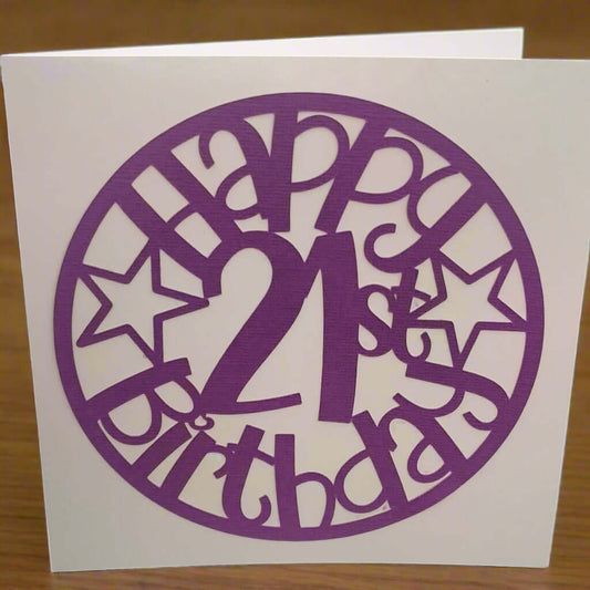 Purple papercut birthday card reading 'Happy 21st Birthday' with star accents, standing upright on a wooden table background.