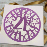 Purple papercut topper reading 'Happy 21st Birthday' in a circular design with stars, displayed on white card with gold ribbon and light wood background.