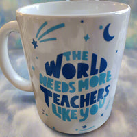 A white ceramic mug with blue text that reads: "THE WORLD NEEDS MORE TEACHERS LIKE YOU." Surrounding the text are blue decorative elements, including stars, a crescent moon, and abstract shapes resembling shooting stars. The mug has a handle on the left and rests on a neutral surface, radiating a sense of appreciation for educators.