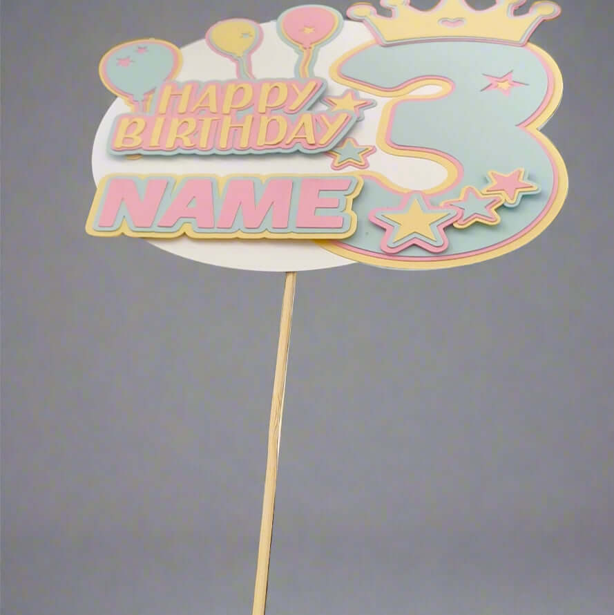 Pastel personalised 3rd birthday cake topper featuring balloons, crown, and stars, displayed upright on a wooden stick against a plain grey background, front-facing view.


