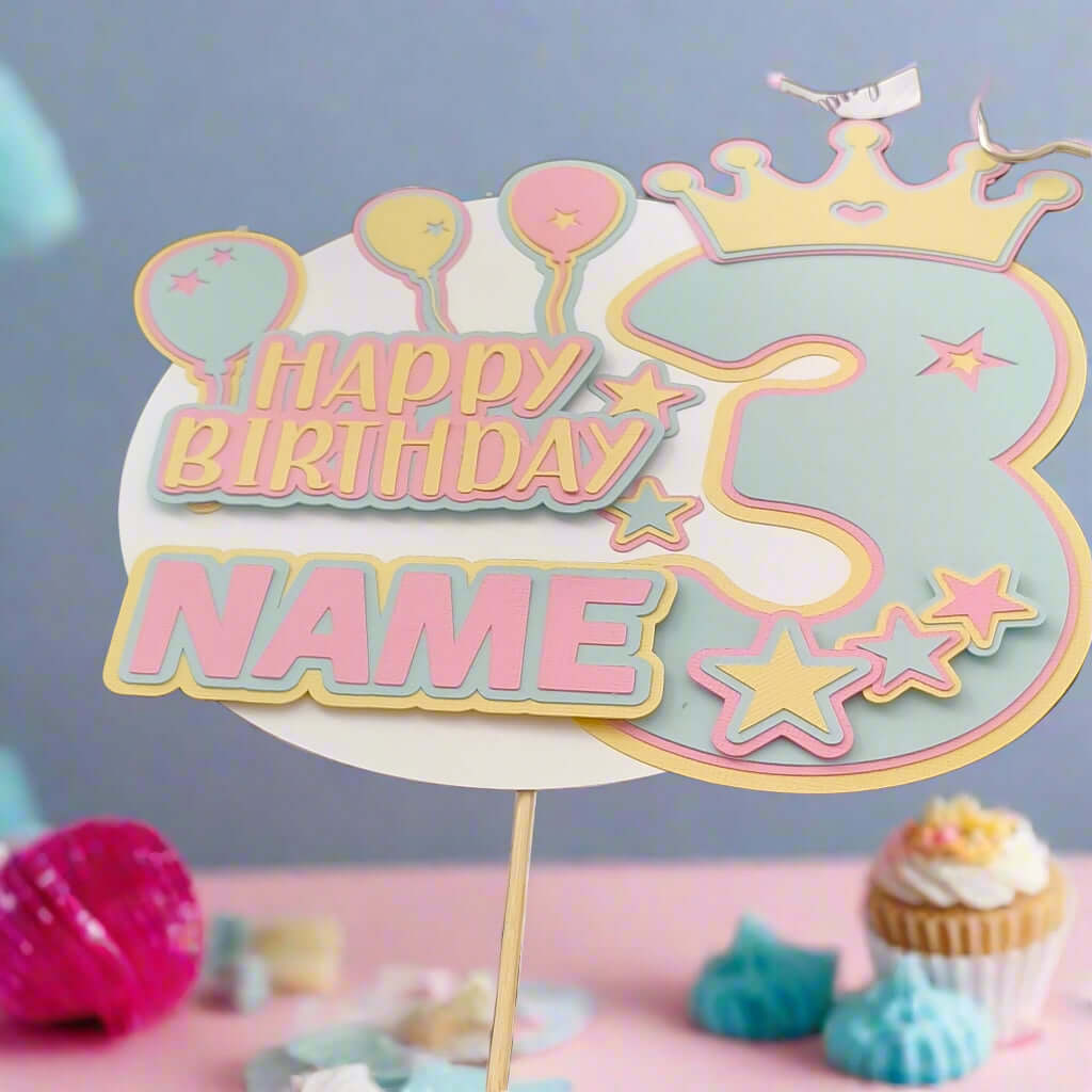 Personalised 3rd birthday cake topper with pastel balloons, stars, and crown, displayed upright on a wooden stick against a party table background with cupcakes and decorations, front-facing angle.

