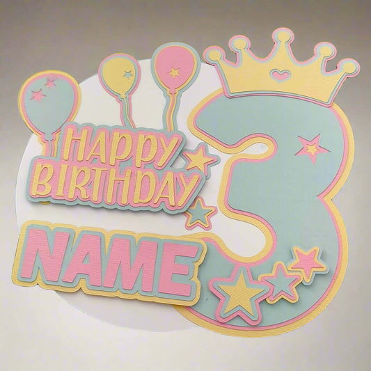 Personalised pastel 3rd birthday cake topper with balloons, stars, and crown, photographed from a direct top-down angle against a softly lit neutral background.

