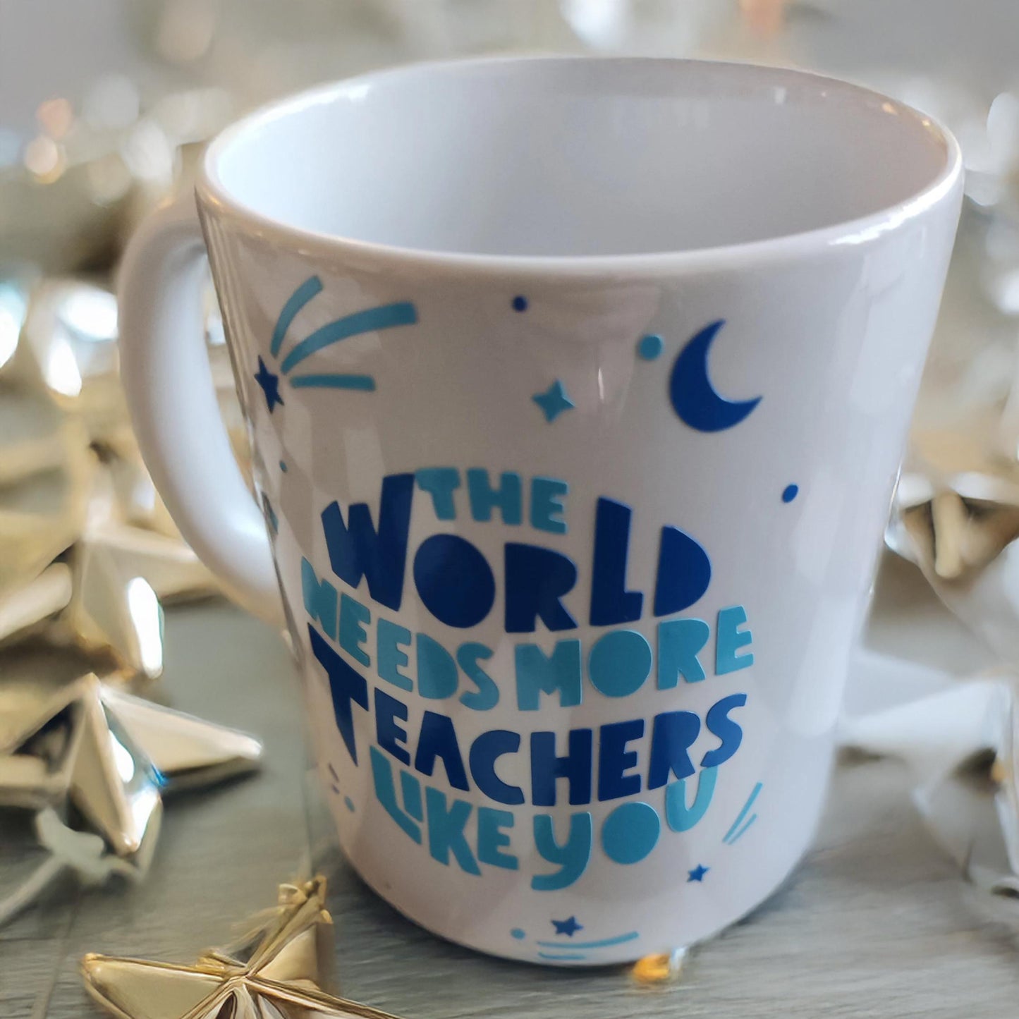A white ceramic mug surrounded by golden star-shaped decorations. The mug features blue text and graphics, including stars, a crescent moon, and dynamic lines. The message on the mug reads: "THE WORLD NEEDS MORE TEACHERS LIKE YOU." The festive and inspiring design celebrates educators and their invaluable contributions.