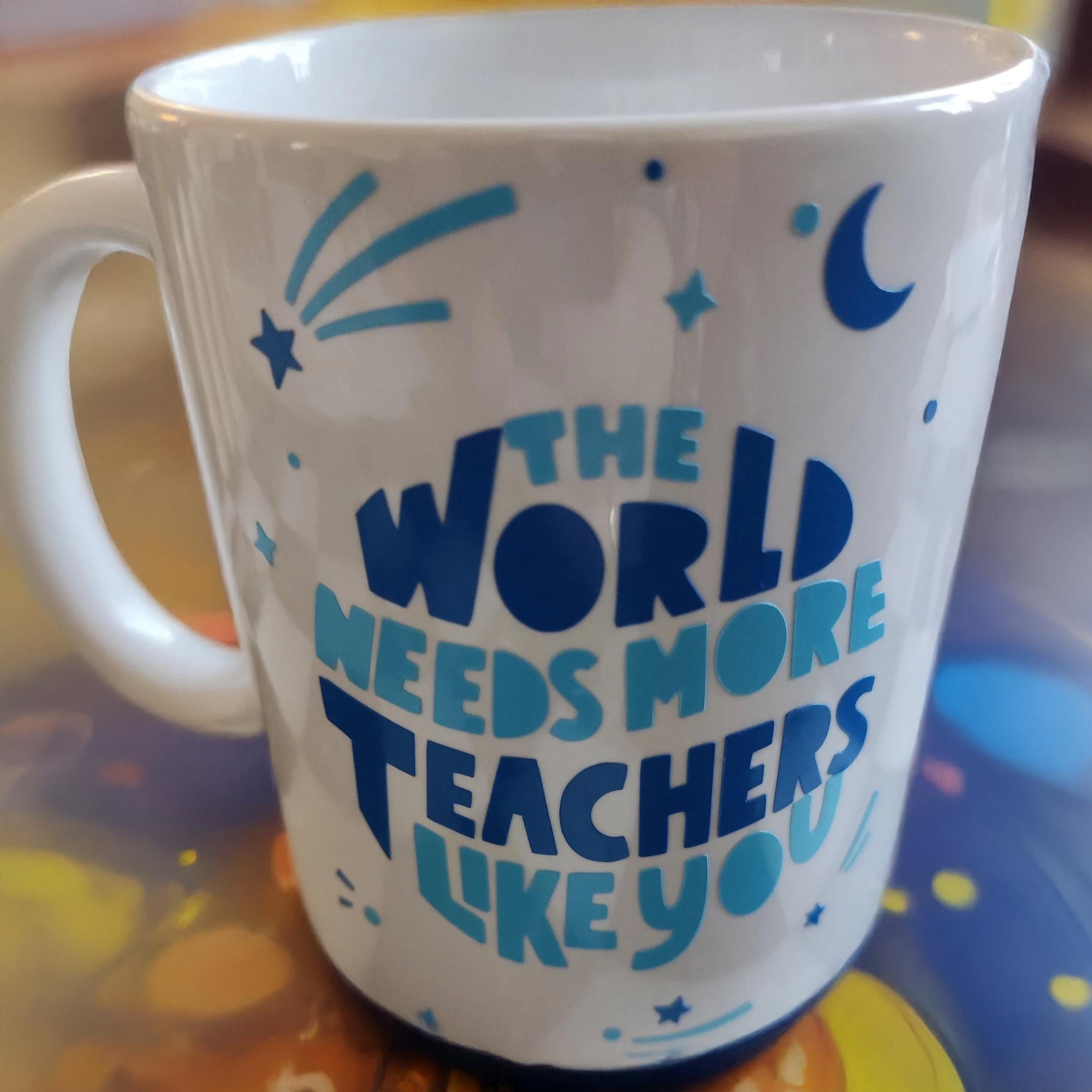 A white ceramic mug featuring blue text that reads, "THE WORLD NEEDS MORE TEACHERS LIKE YOU." The design is adorned with stars, a crescent moon, and shooting stars for a whimsical touch. The mug is displayed on a colourful surface with abstract patterns, creating a vibrant and uplifting presentation.