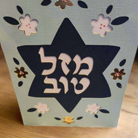 A light blue card stands upright on a wooden surface, featuring a dark blue Star of David cut out in the center. The Hebrew text "מזל טוב" (Mazel Tov) is prominently displayed within the star in white. The card is decorated with cut-out flowers in white, dark blue, and metallic brown.


