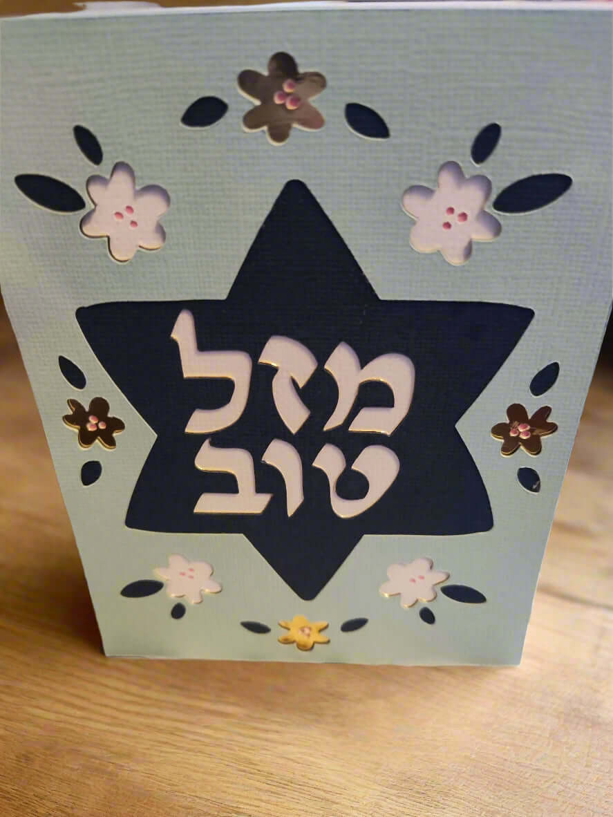 A light blue card stands upright on a wooden surface, featuring a dark blue Star of David cut out in the center. The Hebrew text "מזל טוב" (Mazel Tov) is prominently displayed within the star in white. The card is decorated with cut-out flowers in white, dark blue, and metallic brown.


