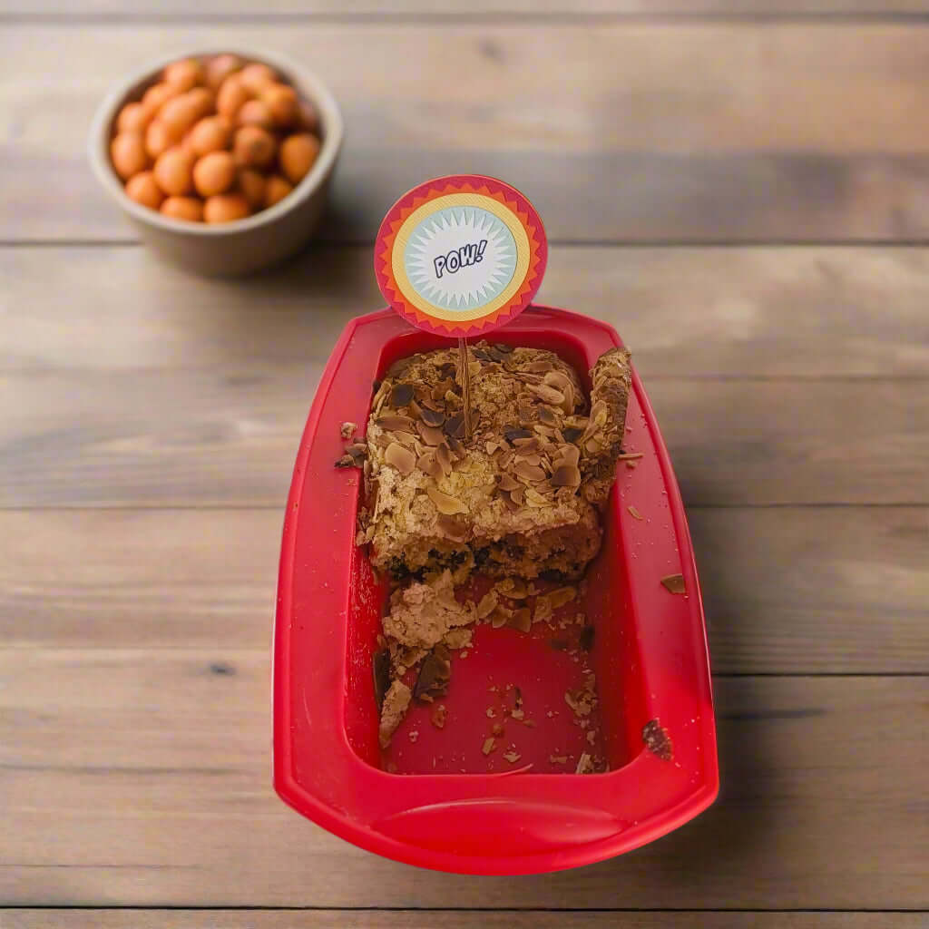 Top-down angle of superhero-style 'POW!' cake topper inserted into an almond loaf cake in a red baking tray, perfect for birthday party decor.