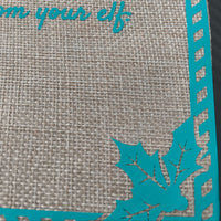 Close-up of a burlap fabric gift bag showing part of the turquoise vinyl text 'from your elf', a striped border, and a decorative holly leaf design in the bottom corner.