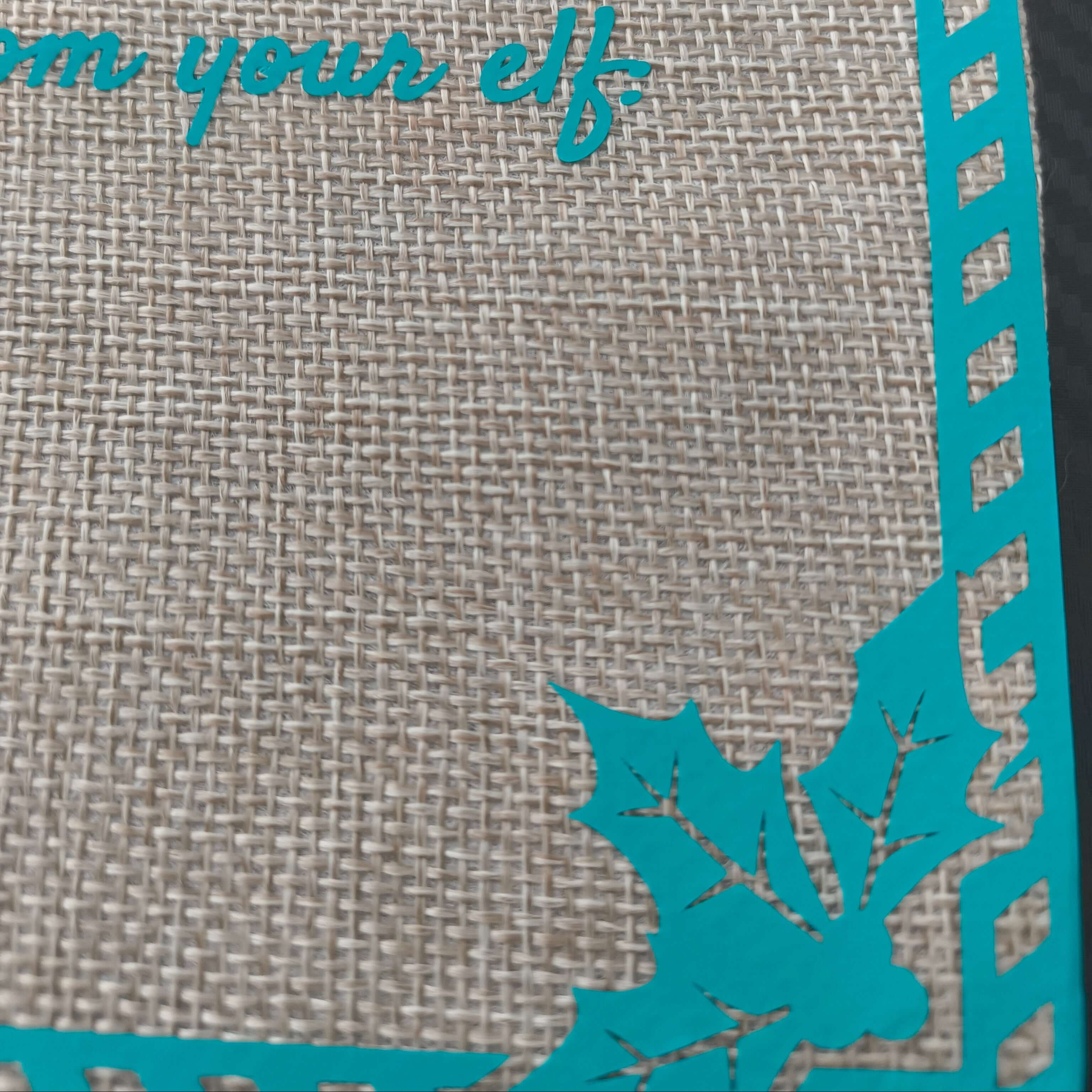 Close-up of a burlap fabric gift bag showing part of the turquoise vinyl text 'from your elf', a striped border, and a decorative holly leaf design in the bottom corner.
