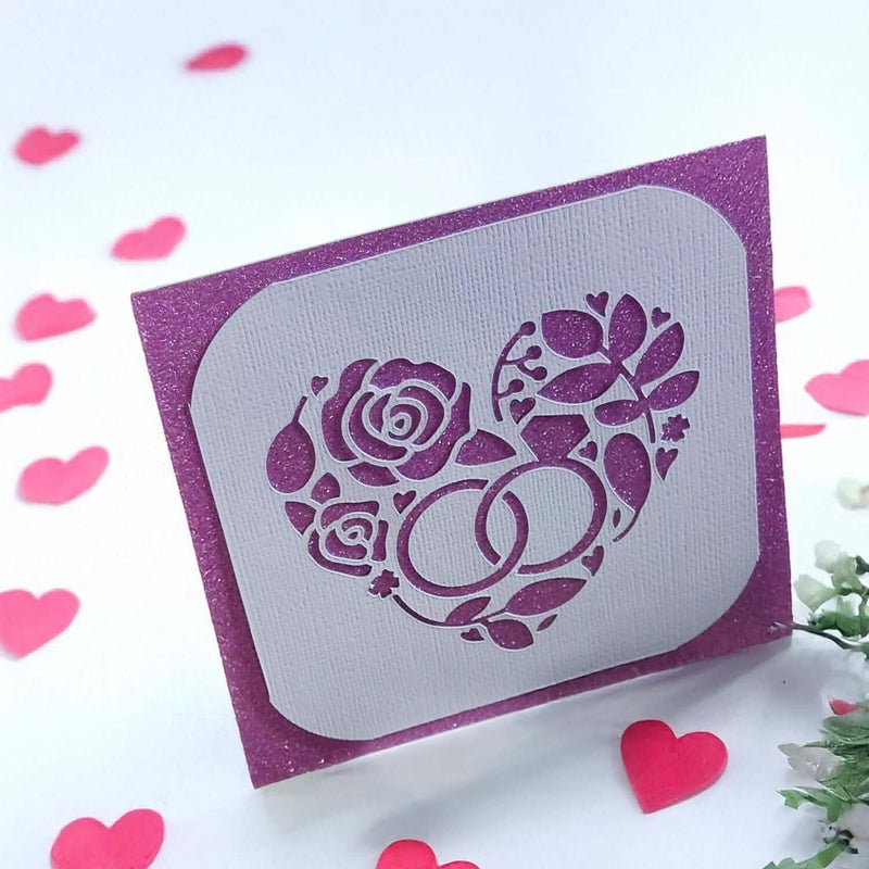 Handmade wedding or anniversary card with a sparkly purple background and a white die-cut floral heart design with wedding rings.


