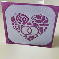 Handmade wedding card with a sparkly purple background and a white die-cut floral heart design with intertwined wedding rings and roses.


