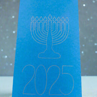 A bright blue Hanukkah card featuring a white line illustration of a menorah with nine candles above the year '2025'. The card stands upright against a soft grey background with scattered white dots resembling falling snow or festive lights, creating a peaceful and celebratory atmosphere.