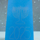 A bright blue Hanukkah card featuring a white line illustration of a menorah with nine candles above the year '2025'. The card stands upright against a soft grey background with scattered white dots resembling falling snow or festive lights, creating a peaceful and celebratory atmosphere.