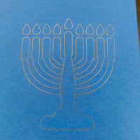 A close-up of a minimalist white line illustration of a menorah with nine candles, finely etched on bright blue card. The design is clean and modern, set against the smooth texture of the card, symbolising Hanukkah in a simple yet elegant style.