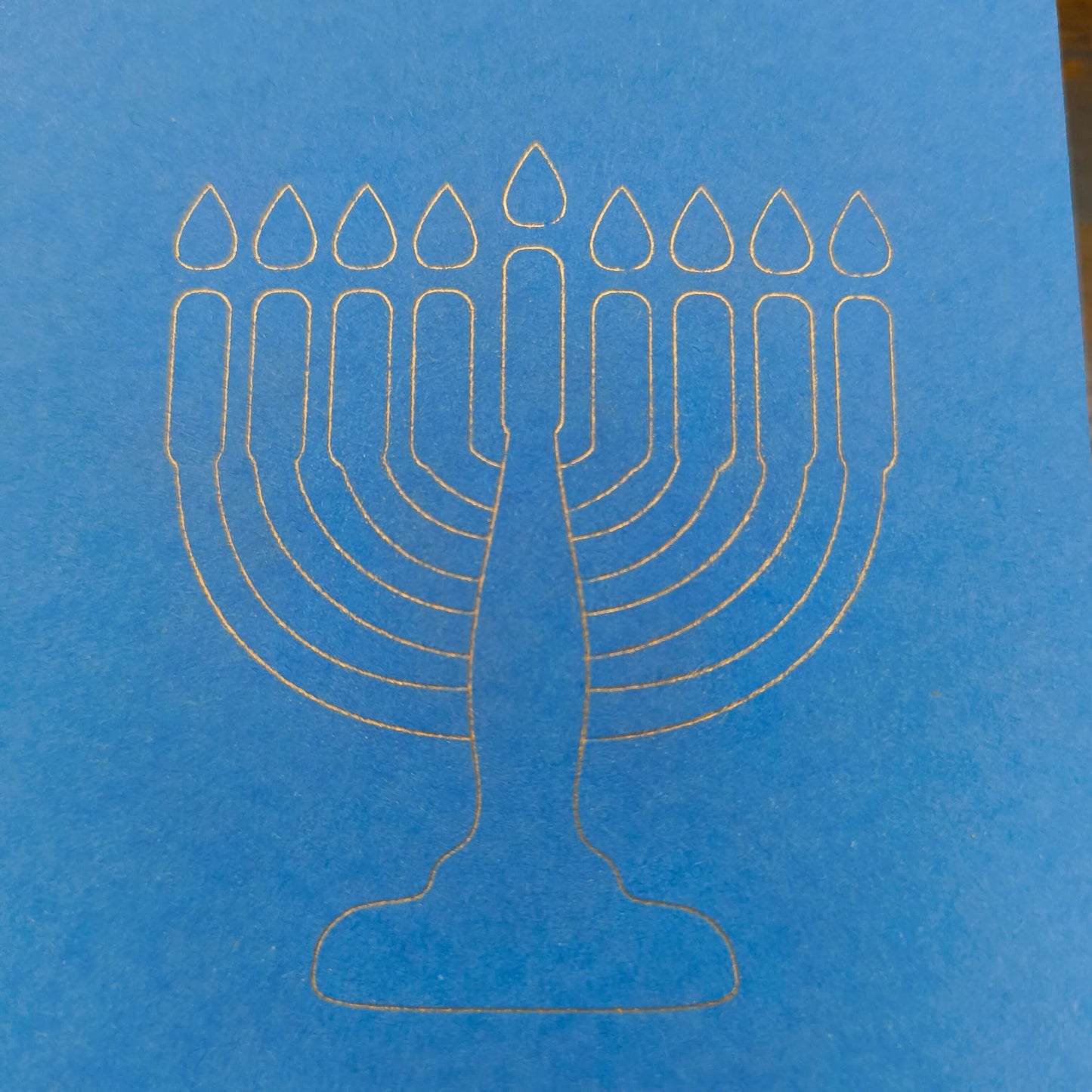 A close-up of a minimalist white line illustration of a menorah with nine candles, finely etched on bright blue card. The design is clean and modern, set against the smooth texture of the card, symbolising Hanukkah in a simple yet elegant style.