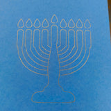 A close-up of a minimalist white line illustration of a menorah with nine candles, finely etched on bright blue card. The design is clean and modern, set against the smooth texture of the card, symbolising Hanukkah in a simple yet elegant style.