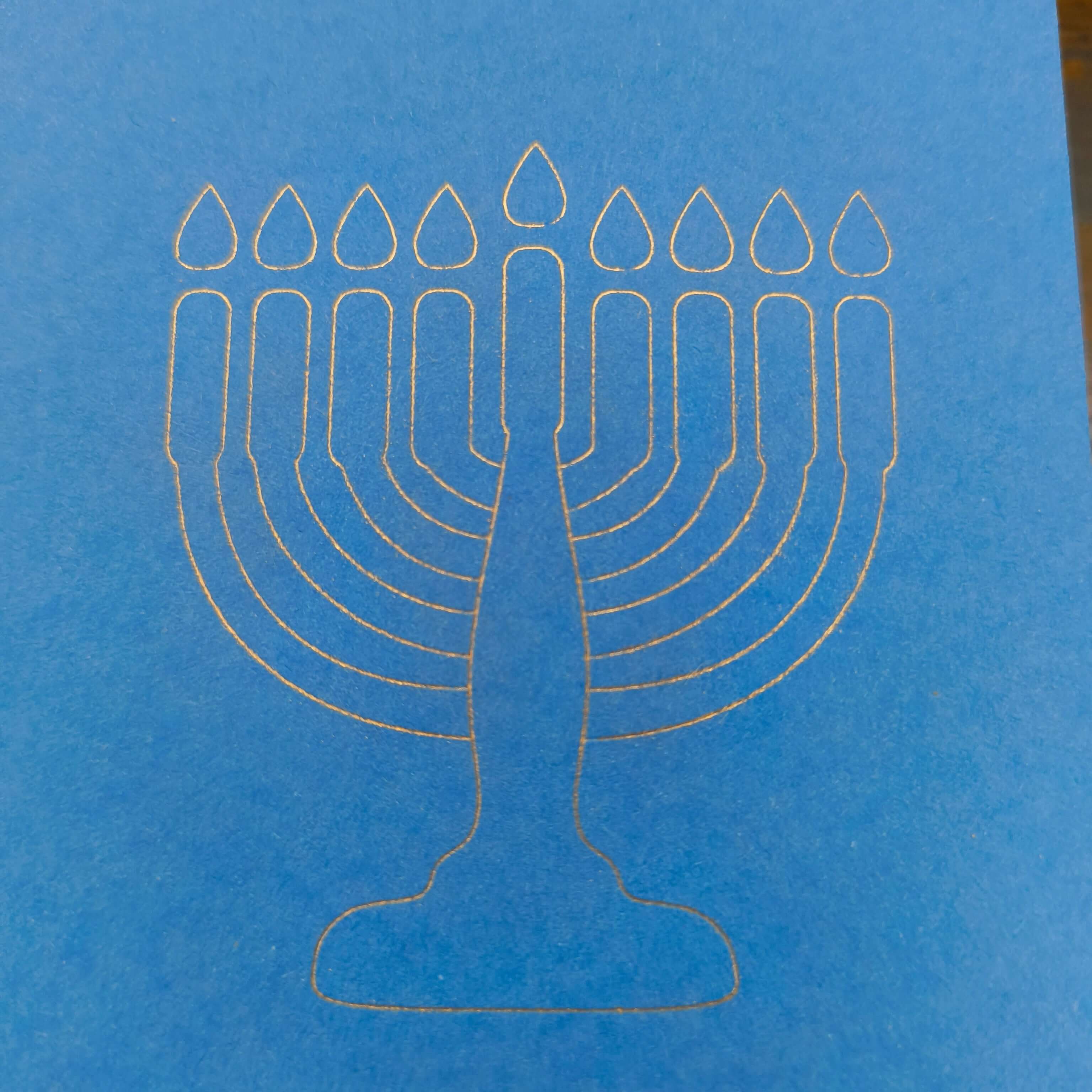 A close-up of a minimalist white line illustration of a menorah with nine candles, finely etched on bright blue card. The design is clean and modern, set against the smooth texture of the card, symbolising Hanukkah in a simple yet elegant style.
