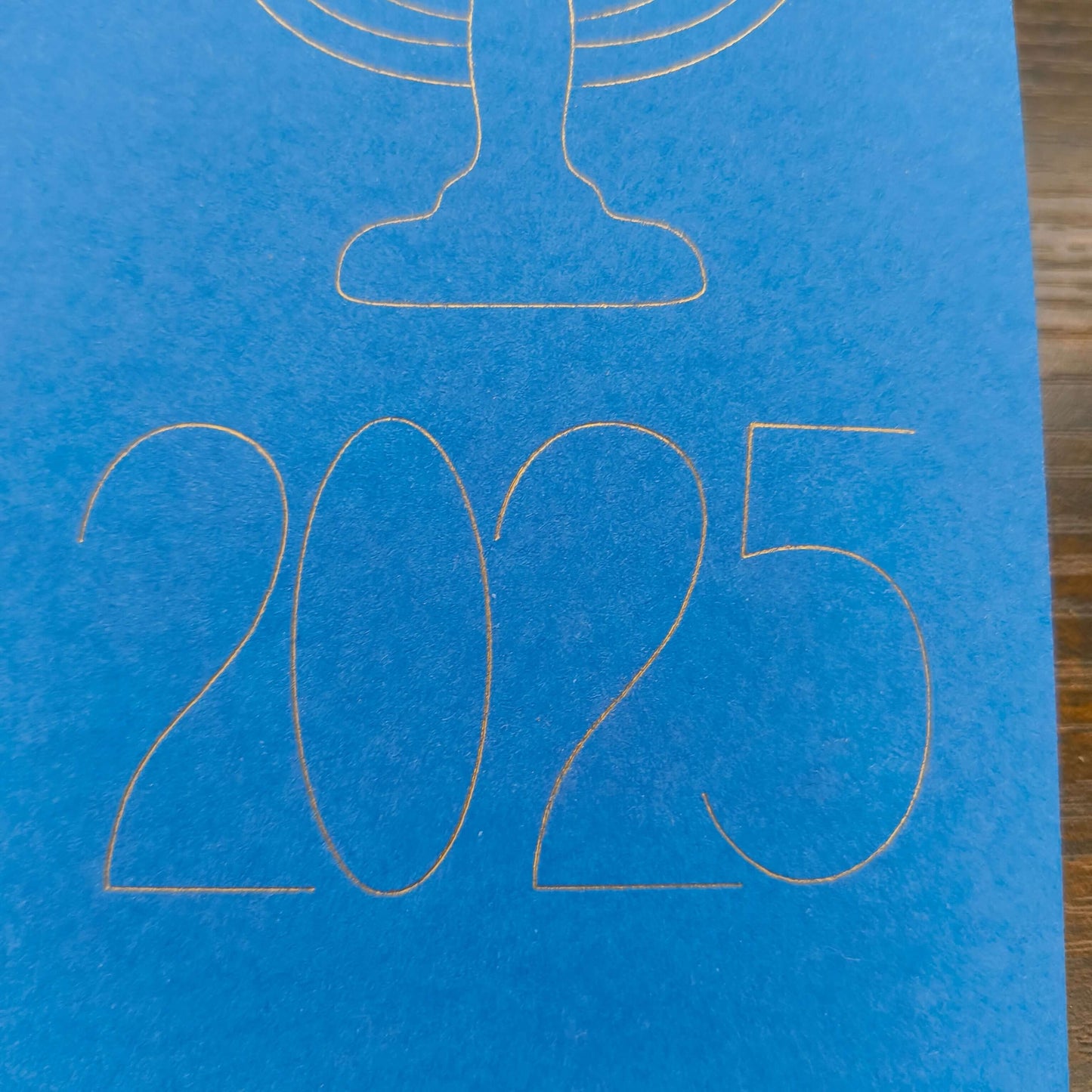 A close-up of the year '2025' finely outlined in gold on a bright blue card. The elegant, modern typography sits beneath the base of a menorah design, part of a minimalist Hanukkah-themed card.