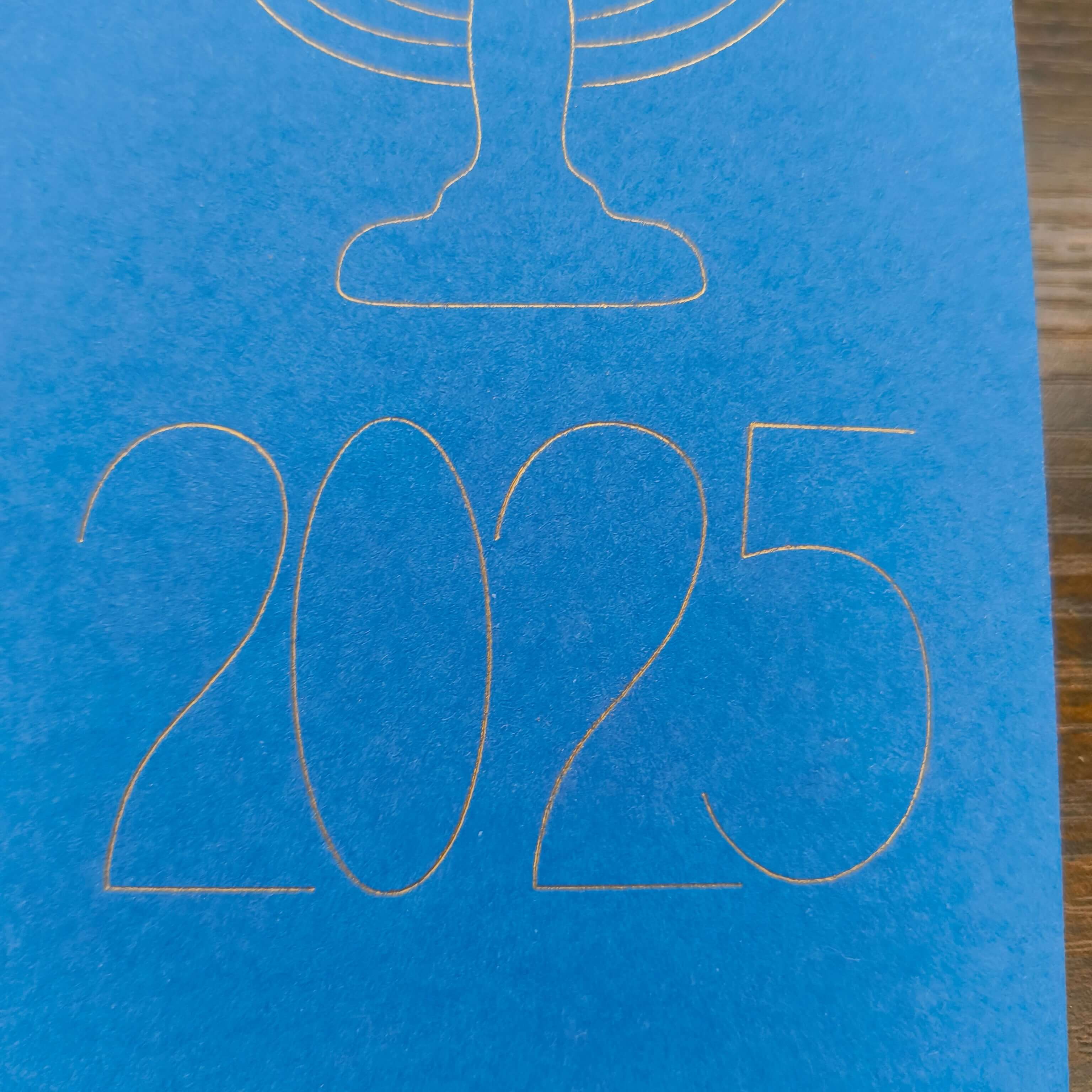 A close-up of the year '2025' finely outlined in gold on a bright blue card. The elegant, modern typography sits beneath the base of a menorah design, part of a minimalist Hanukkah-themed card.