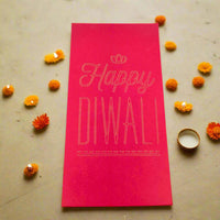 Red Diwali money holder envelope with gold foil-style text reading 'Happy Diwali,' surrounded by marigold flowers and a small tealight candle on a festive stone surface.