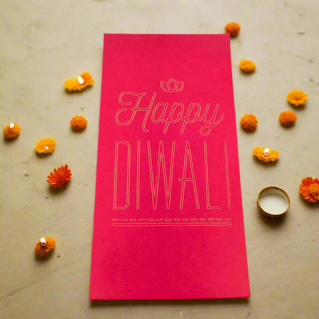 Red Diwali money holder envelope with gold foil-style text reading 'Happy Diwali,' surrounded by marigold flowers and a small tealight candle on a festive stone surface.