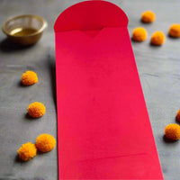 Open red Diwali money envelope displayed on a dark grey festive background, surrounded by small orange marigold flowers and a brass tealight holder.