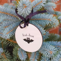 Trick or Treat Halloween bauble with bat design hanging on a pine tree adorned with black ribbon.