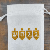 White drawstring hessian gift bag with four gold glitter dreidels printed across the front. Each dreidel features a Hebrew letter: ש (Shin), ה (Hey), ג (Gimel), and נ (Nun), symbolising the Hanukkah saying, "A great miracle happened there." The bag is photographed from above on a rustic wooden surface.
