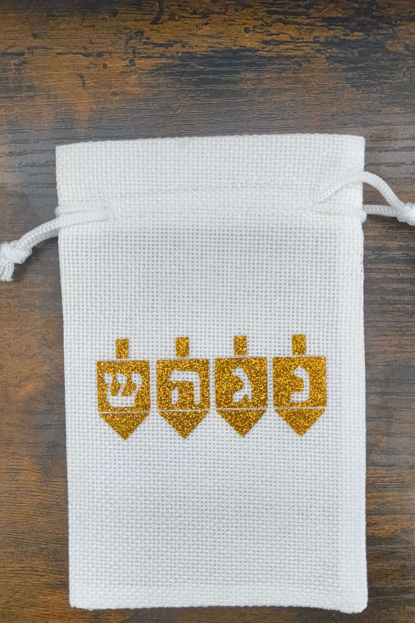 White drawstring hessian gift bag with four gold glitter dreidels printed across the front. Each dreidel features a Hebrew letter: ש (Shin), ה (Hey), ג (Gimel), and נ (Nun), symbolising the Hanukkah saying, "A great miracle happened there." The bag is photographed from above on a rustic wooden surface.
