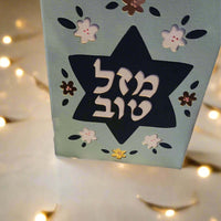 A light blue card stands upright, featuring a dark blue Star of David cut out in the center. The Hebrew text "מזל טוב" (Mazel Tov) is prominently displayed within the star in white. The card is decorated with cut-out flowers in white, dark blue, and metallic brown. The background is blurred with warm yellow lights.