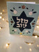 A light blue card stands upright, featuring a dark blue Star of David cut out in the center. The Hebrew text 