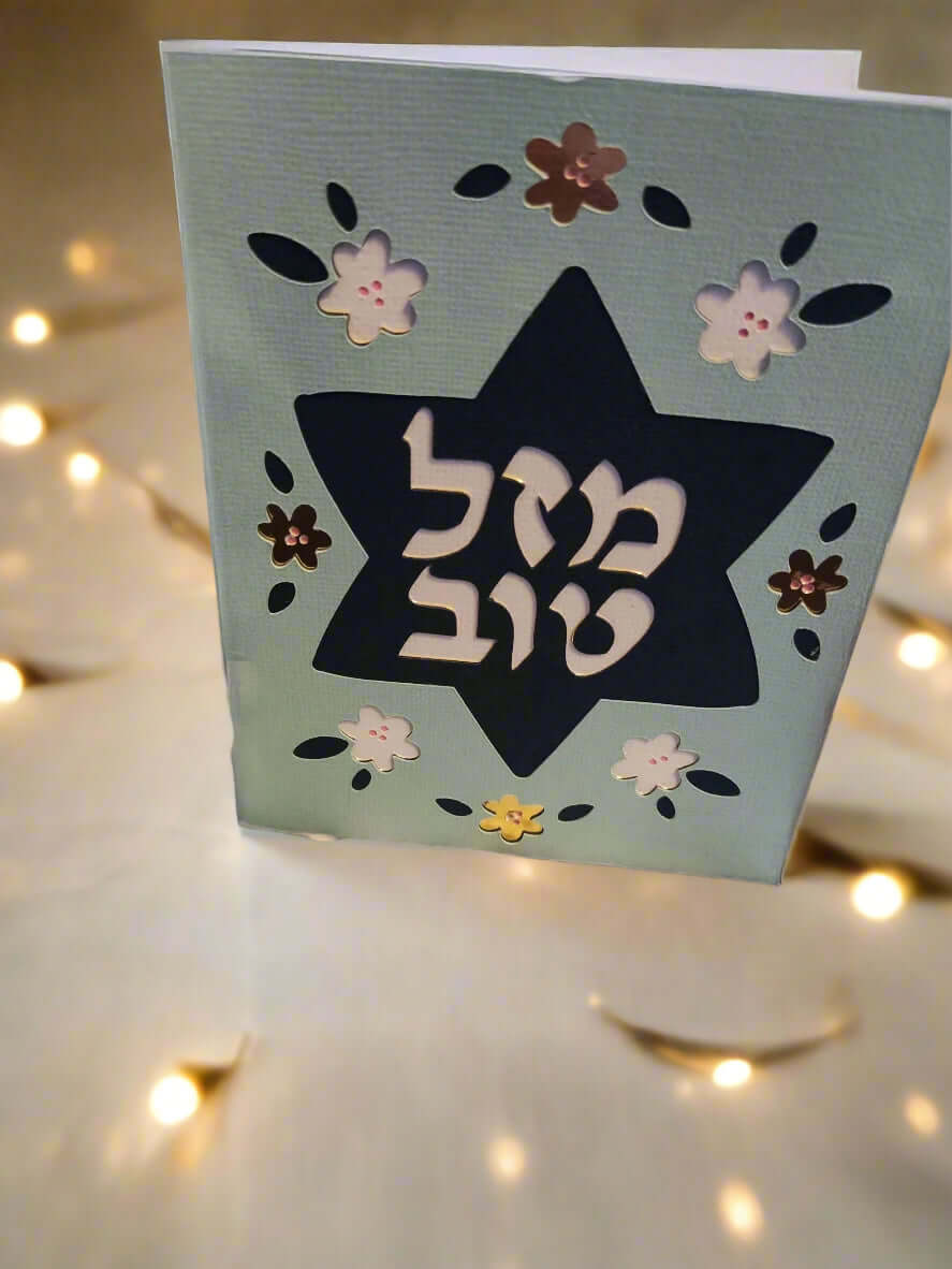 A light blue card stands upright, featuring a dark blue Star of David cut out in the center. The Hebrew text "מזל טוב" (Mazel Tov) is prominently displayed within the star in white. The card is decorated with cut-out flowers in white, dark blue, and metallic brown. The background is blurred with warm yellow lights.