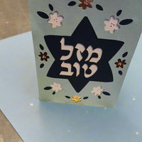 A light blue card stands upright on a light blue surface, featuring a dark blue Star of David cut out in the center. The Hebrew text "מזל טוב" (Mazel Tov) is prominently displayed within the star in white. The card is decorated with cut-out flowers in white, dark blue, and metallic brown.
