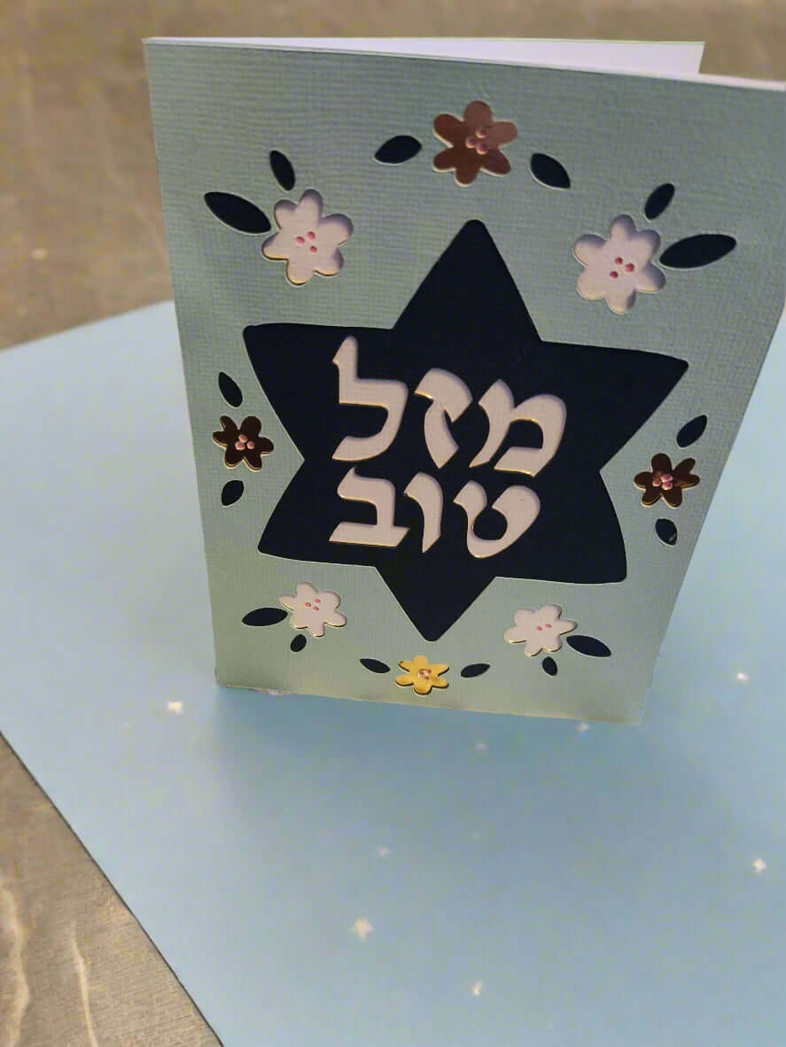 A light blue card stands upright on a light blue surface, featuring a dark blue Star of David cut out in the center. The Hebrew text "מזל טוב" (Mazel Tov) is prominently displayed within the star in white. The card is decorated with cut-out flowers in white, dark blue, and metallic brown.