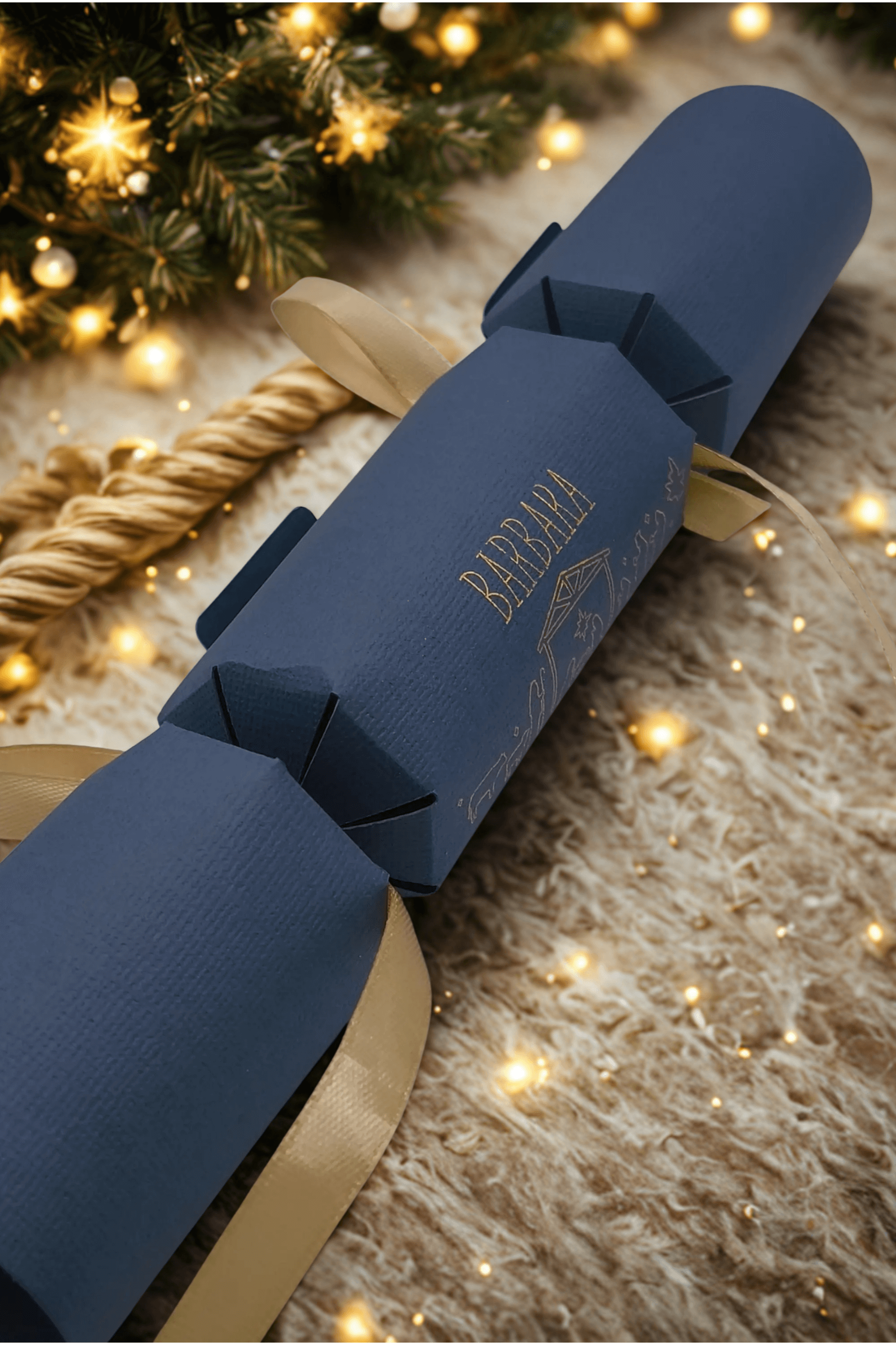 Navy blue personalised Christmas cracker with gold ribbon, displayed on a cosy fur rug with festive tree lights twinkling in the background.