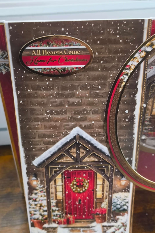 Close-up of Christmas card sentiment reading “All Hearts Come Home for Christmas” with gold edging and snowfall effect.