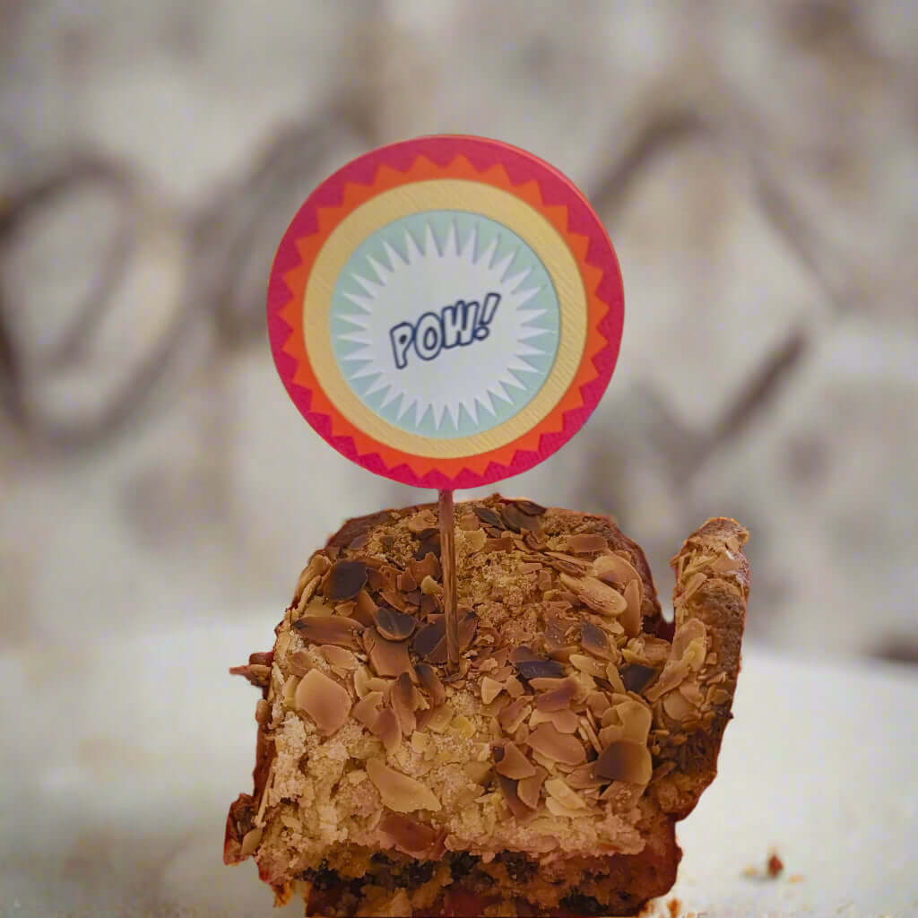 Straight-on view of superhero-themed 'POW!' cake topper inserted into an almond loaf cake, ideal for birthday party decorations.