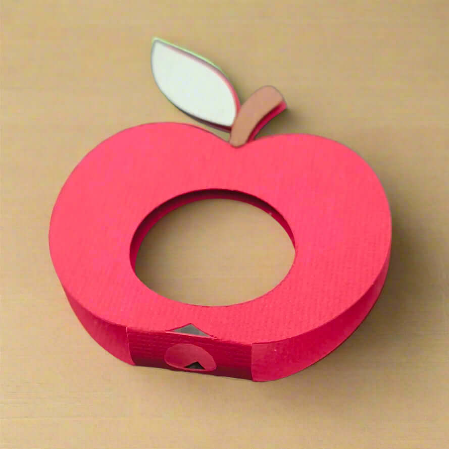 Red apple-shaped handmade lollipop holder made from textured cardstock with a brown stem and green leaf, displayed on a neutral wooden background.