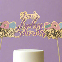 "Gold and purple baby shower cake topper reading 'Lou's Baby Shower', featuring pastel baby icons and floral details. The topper is placed on a white cake against a soft lavender background, creating a clean and elegant product display.