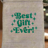 Hessian drawstring gift bag with green retro-style text reading "Best Gift Ever!" placed in front of a softly blurred festive background with warm yellow fairy lights and hints of Christmas décor.