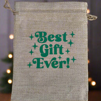 Hessian drawstring gift bag with green retro-style text reading "Best Gift Ever!" displayed against a softly blurred festive background with warm fairy lights and a Christmas tree silhouette.