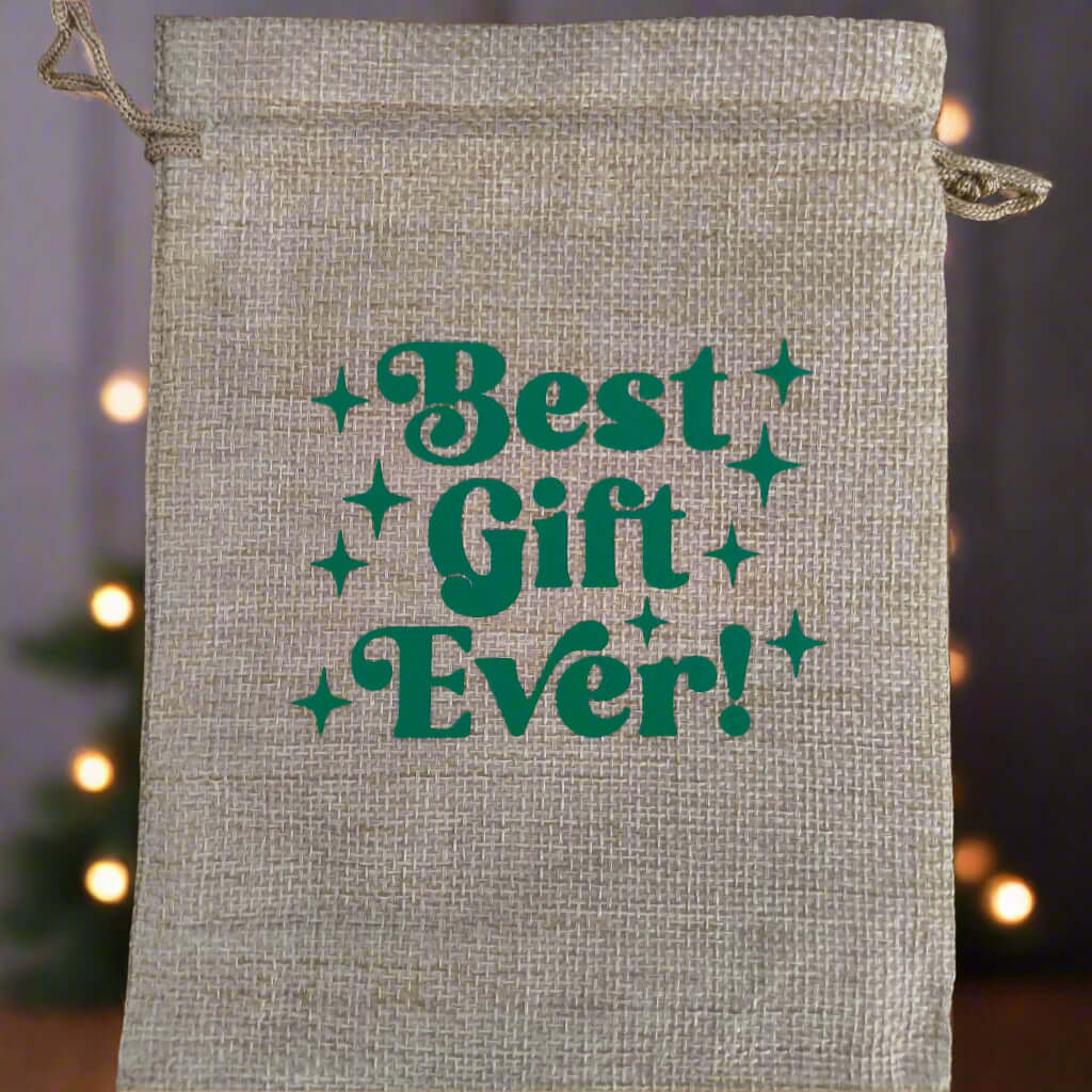 Hessian drawstring gift bag with green retro-style text reading "Best Gift Ever!" displayed against a softly blurred festive background with warm fairy lights and a Christmas tree silhouette.