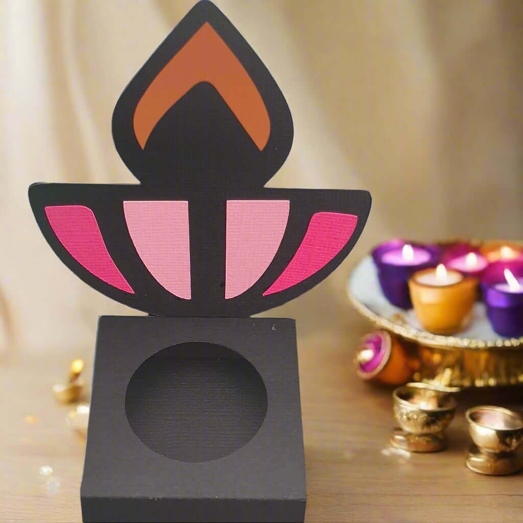 Front-facing image of a black Diwali sweet holder with pink and orange diya design, displayed on a wooden table with festive tealights and gold accents in the background.