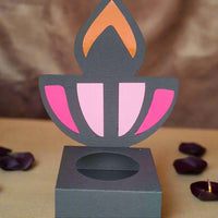 Front-facing image of a handmade black Diwali diya sweet holder with pink and orange flame design, displayed on a beige surface with rose petals and soft festive lighting in the background.