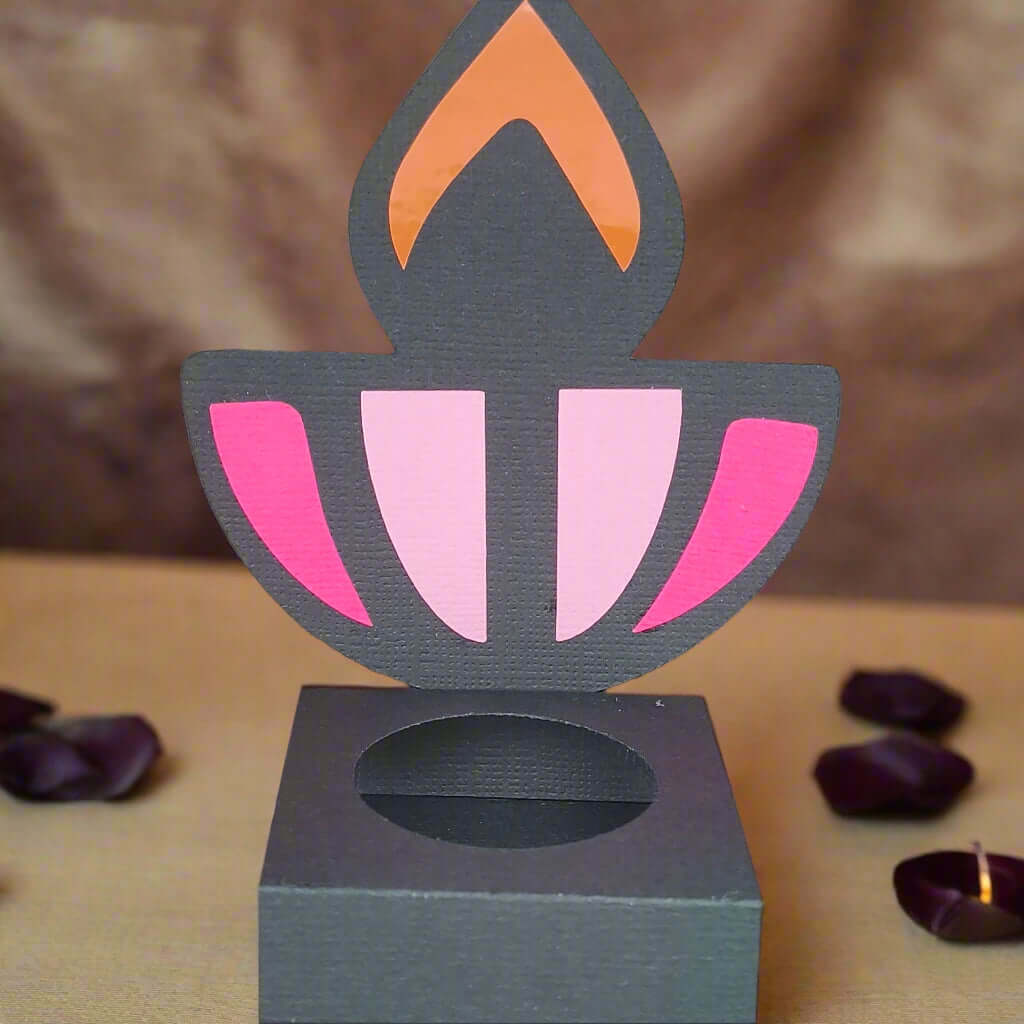 Front-facing image of a handmade black Diwali diya sweet holder with pink and orange flame design, displayed on a beige surface with rose petals and soft festive lighting in the background.