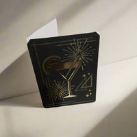 Standing black card with gold foil cocktail glass and fireworks design, shown half open against a neutral background.